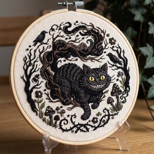 Op de afbeelding: Geborduurd kunstwerk in een houten hoepel met een grijnzende zwarte kat, omringd door donkere takken, paddenstoelen en twee zwarte vogels. De kat heeft gele ogen en een gestreept lichaam. De achtergrond is een lichtbeige stof.