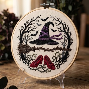 Könnte beinhalten: Gesticktes Kunstwerk in einem Holzrahmen mit Hexenhut, Besen und roten Schuhen. Der schwarze Hut hat lila Akzente, und Fledermäuse fliegen darum herum. Das Design ist von schwarzen Bäumen und grünen Ranken umgeben, was ein Halloween-Thema ergibt.
