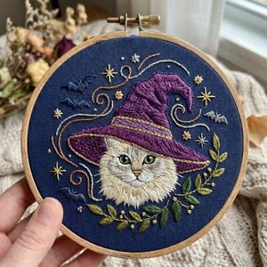 Könnte beinhalten: Gesticktes Kunstwerk mit einer Katze, die einen lila Hexenhut trägt, vor einem dunkelblauen Hintergrund. Das Design umfasst Sterne, Fledermäuse und einen grünen Blätterkranz, alles in einem hölzernen Stickrahmen. Die Katze hat grüne Augen.