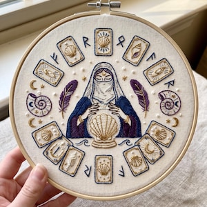 May include: An embroidery hoop containing a white fabric panel with a central figure wearing a head covering and dark blue robes, gazing at a large golden seashell. The design includes surrounding tarot-style cards, seashells, feathers, and runes.