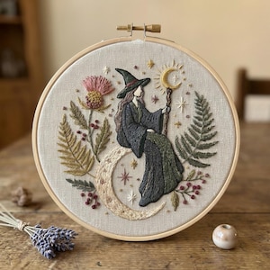 Könnte beinhalten: Gesticktes Kunstwerk in einem Holzreifen. Das Design zeigt eine Hexe in einem schwarzen Kleid und Hut, die auf einem Halbmond sitzt und einen Stab mit einem Halbmond an der Spitze hält. Der Hintergrund umfasst Sterne, Farne und eine Distel.