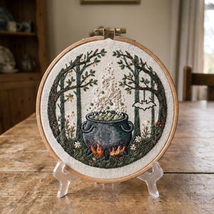 Op de afbeelding: Geborduurd kunstwerk in een houten hoepel toont een ketel die boven een vuur in een bosscene borrelt. Het kunstwerk toont een zwarte ketel, groene bladeren en een vleermuis. De hoepel wordt op een transparante standaard getoond.