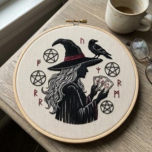 May include: Embroidered artwork in a wooden hoop features a witch silhouette wearing a hat with a raven perched on top. The witch holds cards, surrounded by pentagrams and runes, all in black and red thread on a beige background.