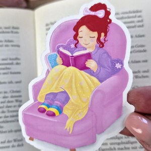 Könnte beinhalten: Aufkleber mit einer Cartoon-Illustration einer Person, die in einem rosa Sessel ein Buch liest. Die Person hat rotes Haar, trägt einen lila Pyjama und eine gelbe Decke. Das Buch ist lila.