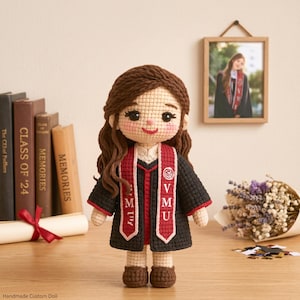 May include: A handmade crochet doll wearing a graduation gown with a red and black design. The doll has brown hair and is standing on a wooden surface. A framed photo and books are in the background.