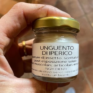 Può includere: Un piccolo barattolo di vetro con coperchio dorato, tenuto in mano. L'etichetta recita "UNGUENTO DI IPERICO" ed elenca gli usi per punture di insetti, scottature solari e dolori muscolari. Ingredienti: olio d'oliva, cera d'api e olio essenziale di lavanda.