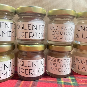 Può includere: Piccoli vasetti di vetro con coperchi dorati, riempiti con varie creme. Ogni vasetto ha un'etichetta con testo in italiano, tra cui "Unguento" e altre parole. I vasetti sono disposti su una superficie a scacchi rossi e verdi.