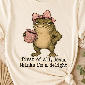 May include: Cream-colored t-shirt featuring a grumpy cartoon frog wearing a pink bow and holding a coffee mug. The frog has a disgruntled expression. The text on the shirt reads "first of all, Jesus thinks I'm a delight."