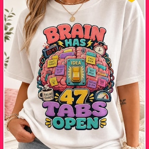 Może przedstawiać: Biała koszulka z kolorowym nadrukiem graficznym. Projekt przedstawia mózg z wieloma zakładkami, zegarem i napisem "BRAIN HAS 47 TABS OPEN". Koszulka zawiera również napis "FUELING CHAOS".