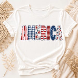 May include: White t-shirt featuring the word "AMERICA" in a patriotic design. The letters are decorated with red and white stripes, blue gingham, red and white checks, blue stars, and floral patterns. The shirt is knotted at the waist.