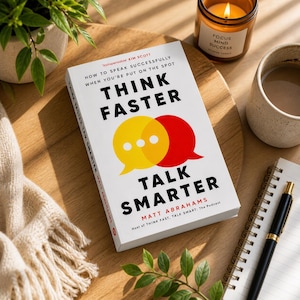 May include: A hardcover book titled "Think Faster, Talk Smarter" with a yellow and red speech bubble design. The book is on a wooden surface, next to a lit candle, a mug, a notebook, and a pen. The book's author is Matt Abrahams.