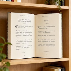 Pode incluir: Um livro aberto com o t&iacute;tulo "Think and Grow Rich" numa prateleira de madeira. As p&aacute;ginas apresentam texto sobre a realiza&ccedil;&atilde;o de objetivos e o poder da cren&ccedil;a. O livro est&aacute; sobre um fundo neutro.