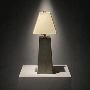 May include: A modern table lamp with a trapezoidal base and a rectangular lampshade. The base is dark gray, and the lampshade is a light beige color. The lamp is placed on a white surface against a dark gray background. The lamp's dimensions are not visible.