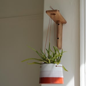 Op de afbeelding: Een hangende bloempot van keramiek, wit met roze en oranje strepen, met een groene plant. De pot hangt aan een wandhouder van licht hout.