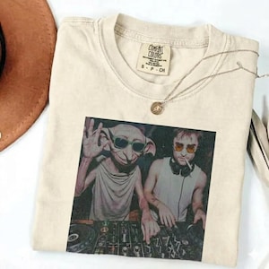 May include: Cream-colored Comfort Colors t-shirt featuring a graphic of two figures wearing sunglasses behind a DJ mixing board. A silver necklace with a pendant is draped on the shirt. The shirt has a Comfort Colors tag.
