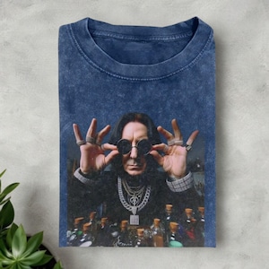 May include: A blue acid wash t-shirt featuring a portrait of a man wearing sunglasses and multiple rings. The man is surrounded by bottles. The shirt has a crew neck and short sleeves.