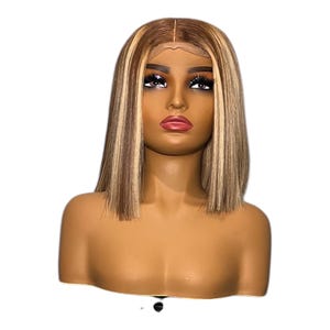 Peut inclure: Une tête de mannequin avec une perruque courte et droite. La perruque a une base marron foncé avec des mèches blondes et une raie au milieu. Le mannequin a un teint hâlé, des lèvres pulpeuses et un maquillage des yeux foncé.