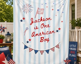 May include: A rectangular fabric backdrop featuring light blue and white vertical stripes. The text reads Jackson is One American Boy in red script. Graphics include a small American flag, red and blue stars, fireworks, and a bunting banner.
