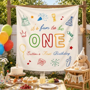 May include: A white fabric backdrop featuring the colourful text it's fun to be ONE and Sutton's First Birthday. The design includes illustrations of a birthday cake, party hat, balloon, gift, and fireworks in blue, red, green, and yellow ink.