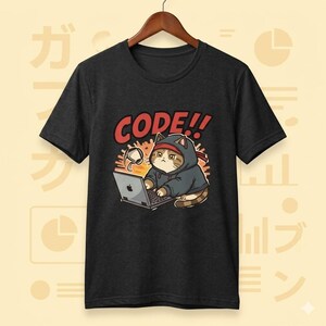 Op de afbeelding: Donkergrijs t-shirt met een cartoonkat met een hoodie die op een laptop werkt. De tekst "CODE!!" staat boven de kat. De kat is bruin en wit met een rode hoofdband. De laptop heeft een Apple-logo.
