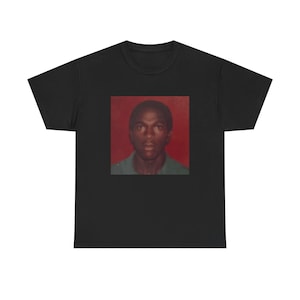 May include: Black t-shirt featuring a portrait of a person with a red background. The portrait is a square image centered on the shirt. The person in the portrait is wearing a green collared shirt.
