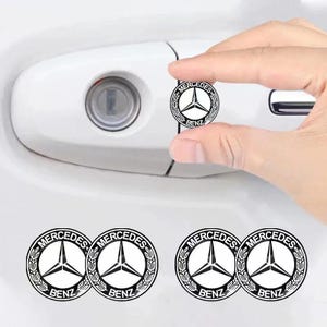 May include: Black and white Mercedes Benz logo stickers. The stickers feature the Mercedes Benz logo with a three-pointed star inside a circle, surrounded by a laurel wreath. The word "BENZ" is printed below the logo. The stickers are shown next to a white car door handle.