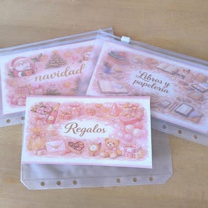 Puede incluir: Tres bolsas de plástico transparente con inserciones decorativas. Las inserciones presentan ilustraciones rosas y texto en español: "navidad", "Libros y papeleria" y "Regalos". Las bolsas están sobre una superficie de madera marrón claro.