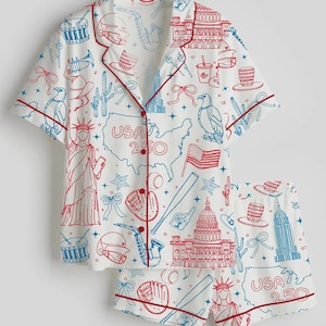 Könnte beinhalten: Weißes Pyjama-Set mit einem rot-blauen USA-Themen-Print. Das kurzärmelige Oberteil und die Shorts zeigen Illustrationen der Freiheitsstatue, des Kapitols und eine Karte der USA. Das Set ist mit roten Paspeln versehen.