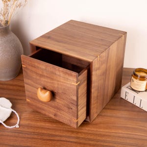 Puede incluir: Una caja de almacenamiento cuadrada hecha de madera de nogal marrón oscuro con un solo cajón extraíble. El frente del cajón presenta un pomo de madera de color claro, liso y redondeado, y uniones de madera clara visibles en las esquinas.