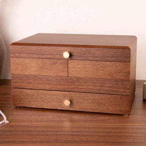 May include: A rectangular wooden jewelry box with a dark brown finish, featuring a top drawer with a single gold knob, two middle drawers, and a bottom drawer with a single gold knob. The box has a smooth, polished surface and clean lines.