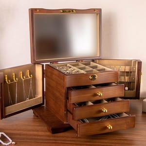 May include: A dark wood jewelry box featuring a hinged lid with an interior mirror, two side panels with gold-toned hooks for necklaces, and three pull-out drawers with gold-toned knobs. The top compartment contains multiple small storage sections.