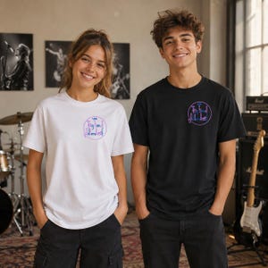 Puede incluir: Una camiseta blanca y una camiseta negra, cada una con un gráfico circular en el pecho izquierdo. El gráfico contiene el texto "The Big Band Fiddlers" en una fuente morada estilizada, encerrado dentro de un fino contorno circular morado en la tela.