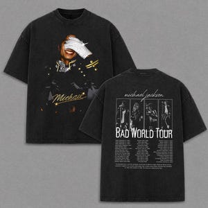 May include: Black t-shirt featuring a graphic of Michael Jackson with a bedazzled glove. The front of the shirt has the name "Michael" in gold script. The back of the shirt has a tour graphic with the text "BAD WORLD TOUR".