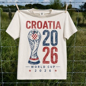 May include: An off-white short-sleeve t-shirt featuring a distressed graphic with the text CROATIA 20 26 WORLD CUP 2026. The design includes a central trophy illustration with a red and white checkered emblem and blue accents.