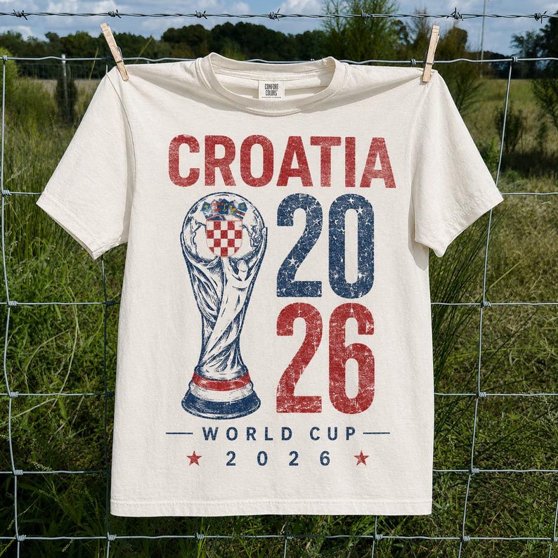 May include: An off-white short-sleeve t-shirt featuring a distressed graphic with the text CROATIA 20 26 WORLD CUP 2026. The design includes a central trophy illustration with a red and white checkered emblem and blue accents.
