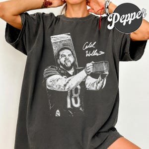 May include: A charcoal gray short-sleeve t-shirt featuring a black and white graphic print of a football player wearing jersey number 18, holding a cheese grater. The graphic includes a handwritten-style signature above the player's image.