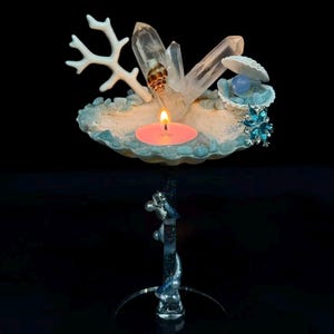 May include: A decorative candle holder featuring a seashell-shaped base with a lit pink candle. The holder is adorned with quartz crystals, a white coral branch, and blue accents. The holder is supported by a clear, ornate stand.