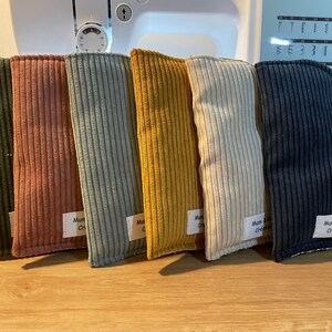 Puede incluir: Seis bolsas de tela de pana en varios colores: verde oliva, coral, azul claro, amarillo mostaza, crema y azul marino. Cada bolsa tiene forma rectangular y una pequeña etiqueta blanca. Las bolsas están dispuestas sobre una superficie de madera.