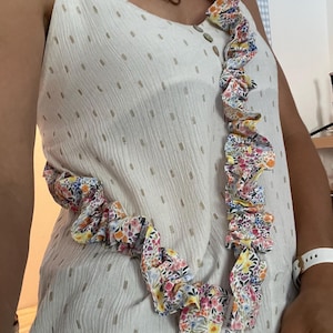 Peut inclure: Un haut sans manches blanc avec un motif de tirets dorés discrets et une fermeture boutonnée, porté avec une longue sangle en tissu à volants ornée d'un imprimé floral coloré dans les tons rose, jaune, bleu et vert, avec une étiquette drapeau.