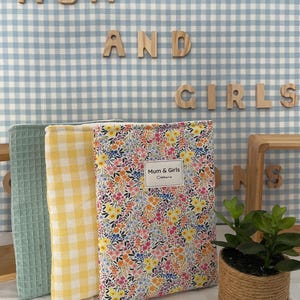 Puede incluir: Tres diarios cubiertos de tela en verde, amarillo a cuadros y estampado floral. El diario floral tiene una etiqueta que dice "Mum & Girls Creations". Letras de madera deletrean "MUM AND GIRLS" sobre un fondo a cuadros azul y blanco. También se ve una pequeña planta en maceta.
