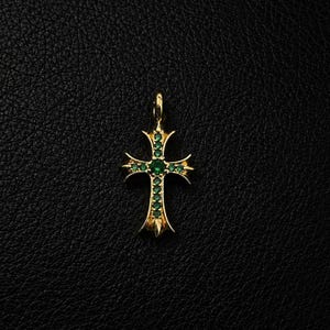 Può includere: Un ciondolo a croce in oro dal design svasato con estremità appuntite. La superficie della croce è incastonata con piccole pietre preziose rotonde di un verde vibrante disposte in un motivo verticale e orizzontale, con una pietra più grande al centro.