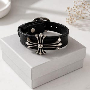 May include: A black leather bracelet featuring a prominent silver-toned metal cross emblem at the centre. The bracelet includes a buckle closure with multiple adjustment holes and a single silver-toned metal rivet securing the leather strap.