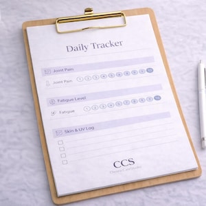 Puede incluir: Un portapapeles de madera con una hoja blanca de 'Daily Tracker', sujeta por un clip dorado. La hoja incluye secciones para dolor articular, nivel de fatiga y registro de piel y UV. Un bolígrafo blanco descansa al lado.