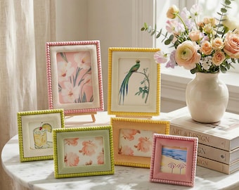 May include: A collection of six rectangular and square picture frames in various pastel colors including pink, yellow, lime green, and peach. Each frame features a beaded border design and contains a different decorative print inside the glass.