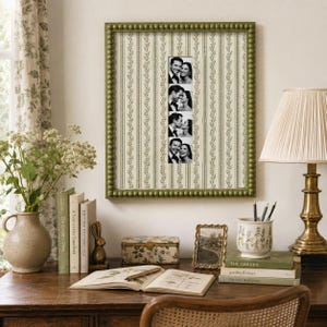 May include: A rectangular wall frame with a beaded olive green border containing a vertical strip of four black and white photographs of a couple, set against a cream-colored background with a repeating green vertical vine and stripe pattern.