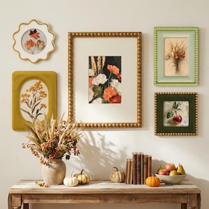 Könnte beinhalten: Eine Galeriewand mit fünf gerahmten botanischen Drucken, darunter Blumen- und Fruchtmotive. Auf einem Holztisch stehen eine Keramikvase mit getrocknetem Weizen und Beeren, drei kleine Kürbisse, alte Bücher sowie eine Schale mit frischem Obst.