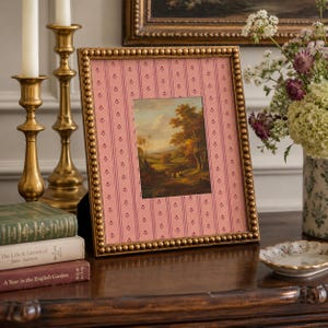 May include: A rectangular picture frame with a metallic gold beaded border. The frame contains a pink mat with a vertical striped pattern featuring small floral motifs. Inside the mat is a landscape painting depicting trees and a path.