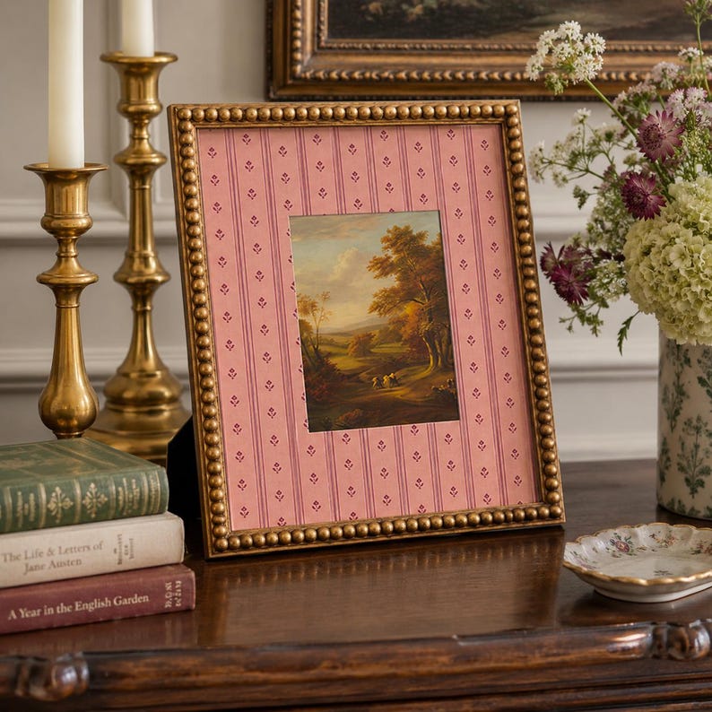 May include: A rectangular picture frame with a metallic gold beaded border. The frame contains a pink mat with a vertical striped pattern featuring small floral motifs. Inside the mat is a landscape painting depicting trees and a path.
