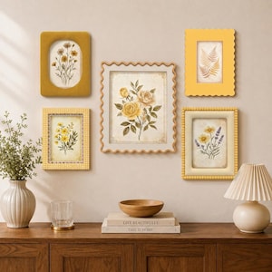 Puede incluir: Una composición de pared con cinco marcos de fotos rectangulares en varios tonos de amarillo, que muestran ilustraciones botánicas de flores y helechos. Los marcos están montados sobre una consola de madera con una lámpara y libros.