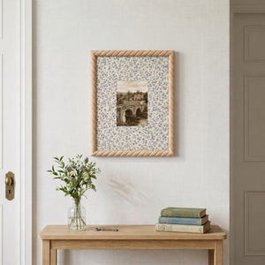 May include: A rectangular picture frame with a light wood finish and a twisted rope texture border. The frame contains a mat with a blue and white floral pattern surrounding a central black and white photograph of a stone bridge and building.